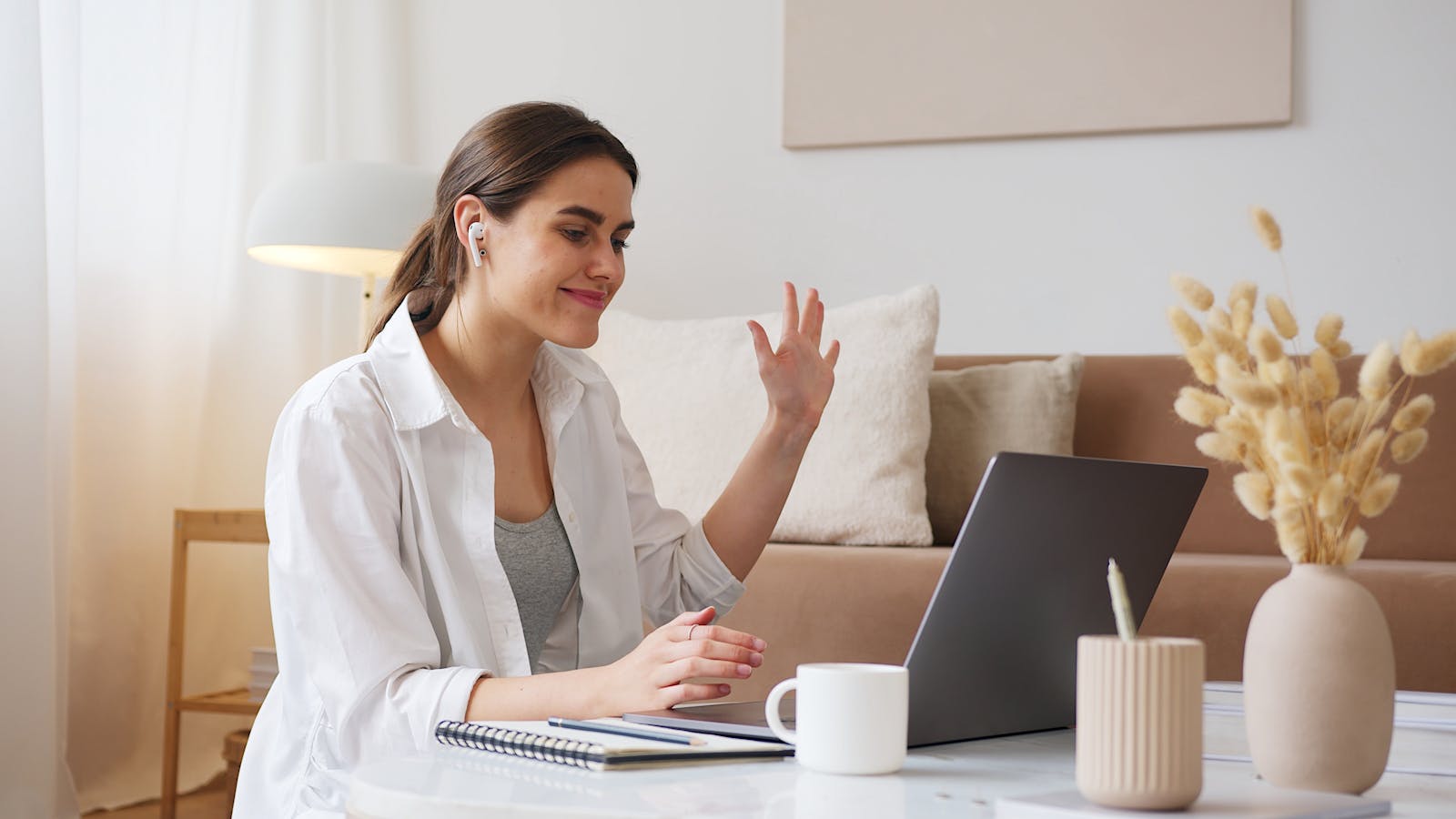 A practitioner in a warm-lit home office on a video call with a patient, laptop open, notebook and mug on the desk